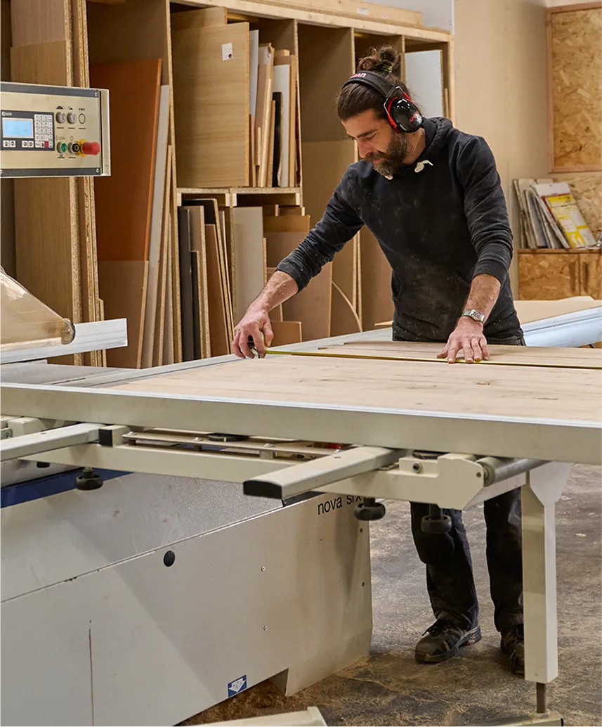 Homme mesurant une planche en bois dans un atelier de menuiserie avec un ruban à mesurer, portant des protections auditives.