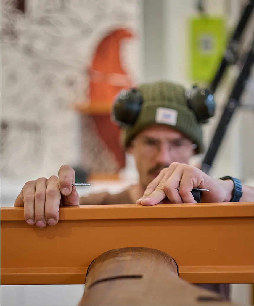 Homme portant un bonnet vert et des protections auditives, tenant une vis au-dessus d’un meuble en bois orange.