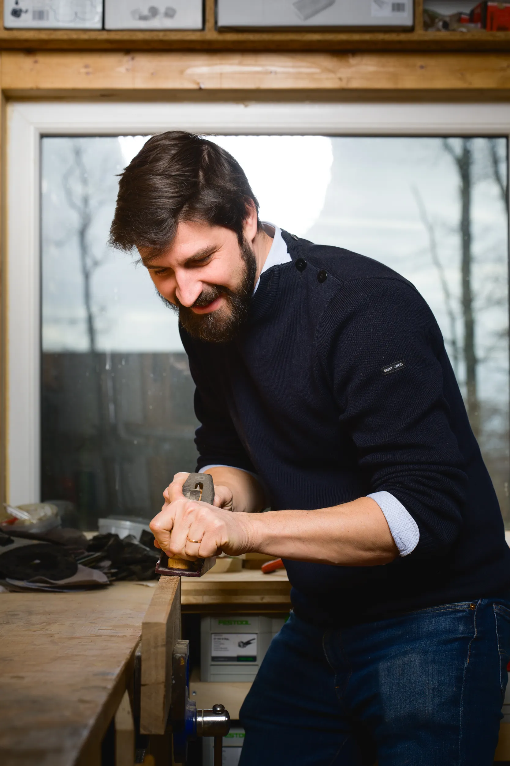 Homme souriant rabotant une planche de bois dans un atelier avec une fenêtre en arrière-plan.